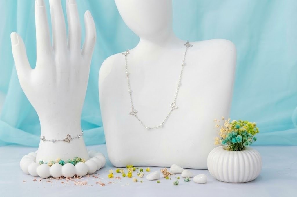 Brazalete y collar plateados con mariposas y perlas sobre una mano y un busto blancos, con flores de colores